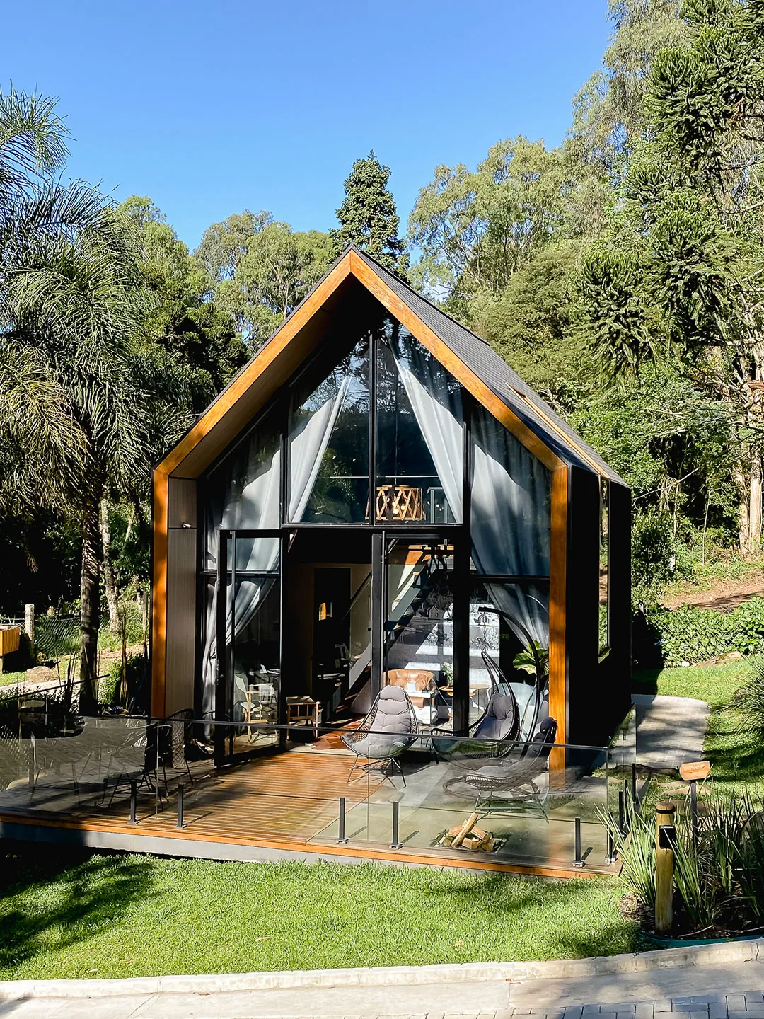 Fachada frontal envidraçada da lake house com cortinas abertas, deck com poltronas e guarda-corpo de vidro, sob céu azul