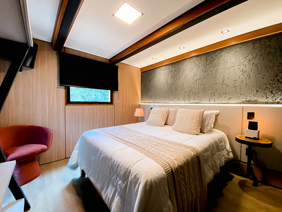 Quarto com cama de casal, parede de textura escura com iluminação indireta e poltrona vermelha