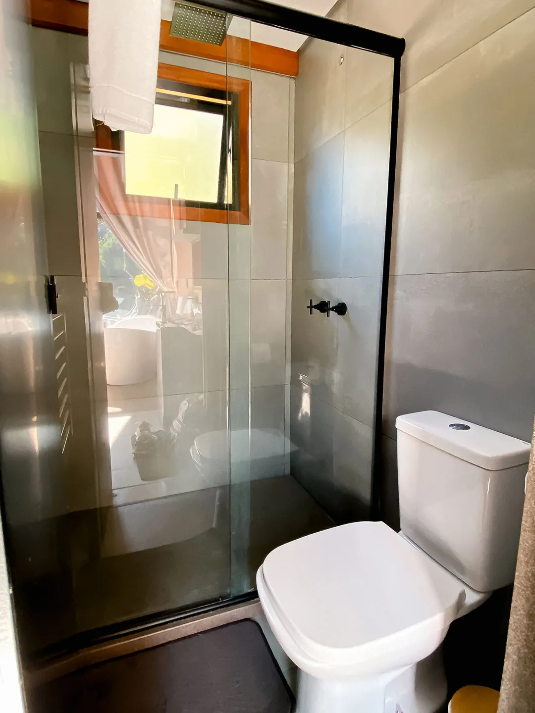 Banheiro com box de vidro, chuveiro e vaso sanitário, com janela de moldura de madeira