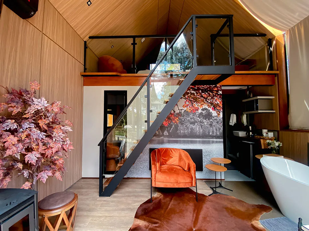 Interior com pé-direito duplo, escada metálica até o mezanino, poltrona laranja, banheira e painel fotográfico de paisagem na parede