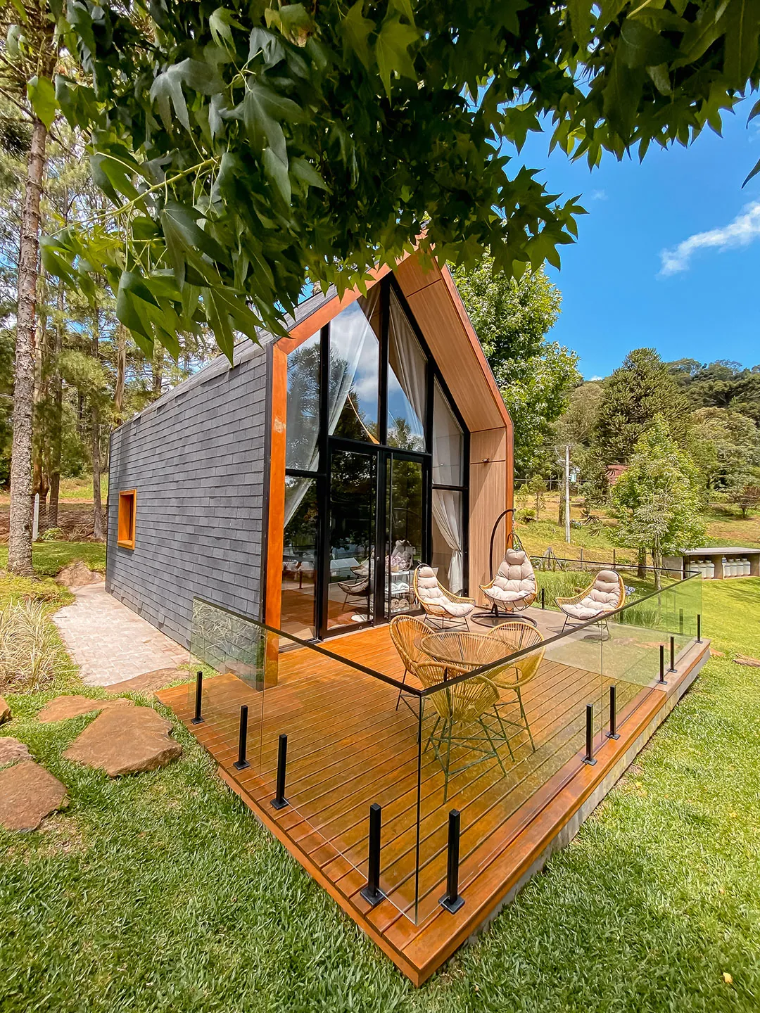 Lateral da lake house com deck de madeira e guarda-corpo de vidro sobre o gramado, sob galhos de árvore
