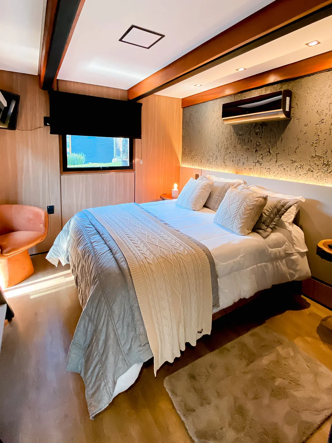 Quarto com cama de casal, manta tricotada, poltrona laranja e ar-condicionado sobre parede texturizada