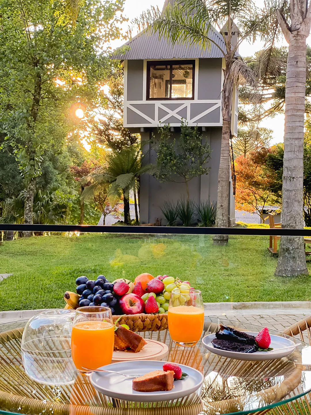Mesa com sucos, frutas e bolo no deck, com gramado e construção de madeira cinza ao fundo contra o sol baixo