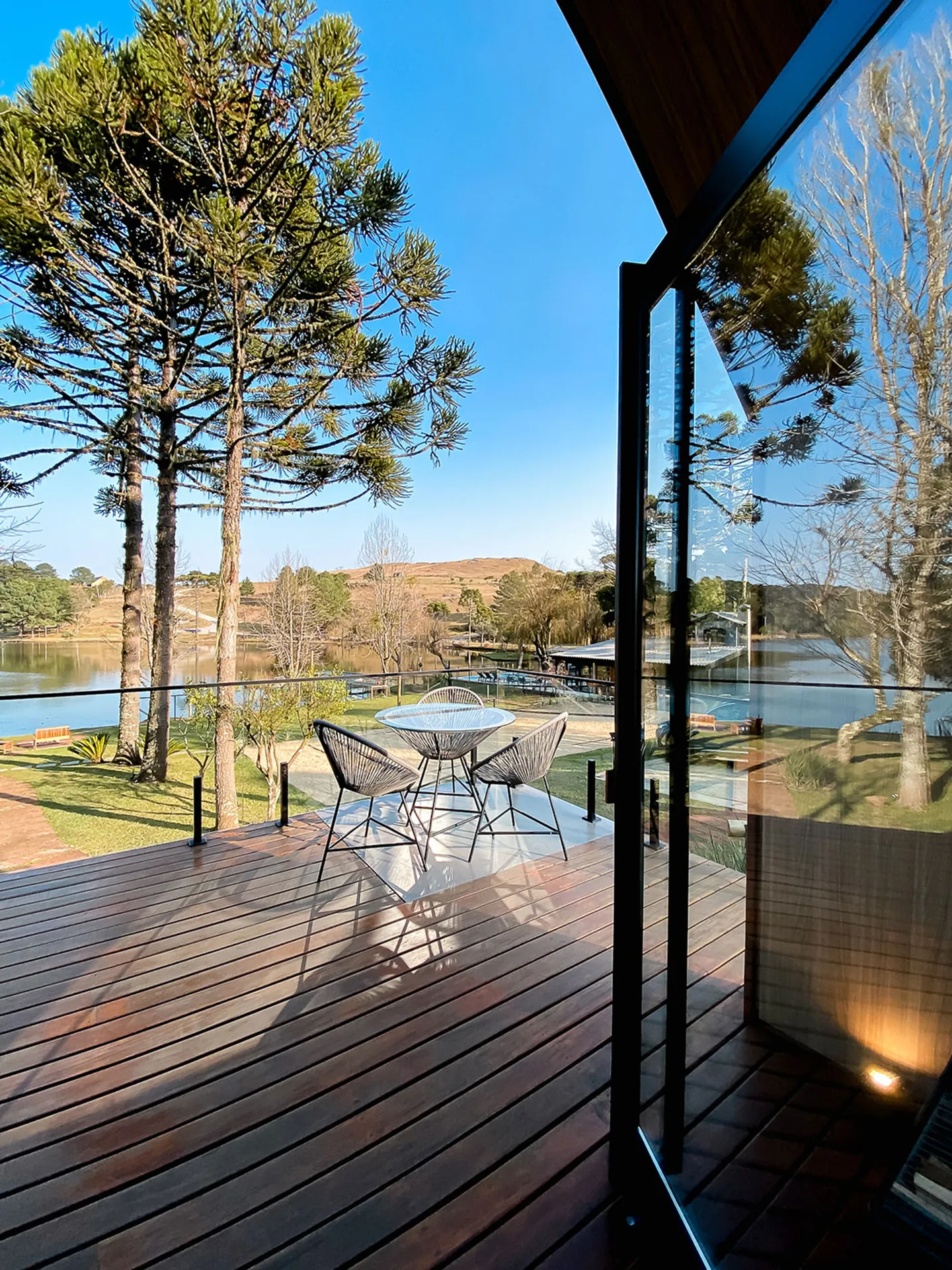 Deck de madeira com mesa e cadeiras de corda, guarda-corpo de vidro e vista para o lago entre araucárias