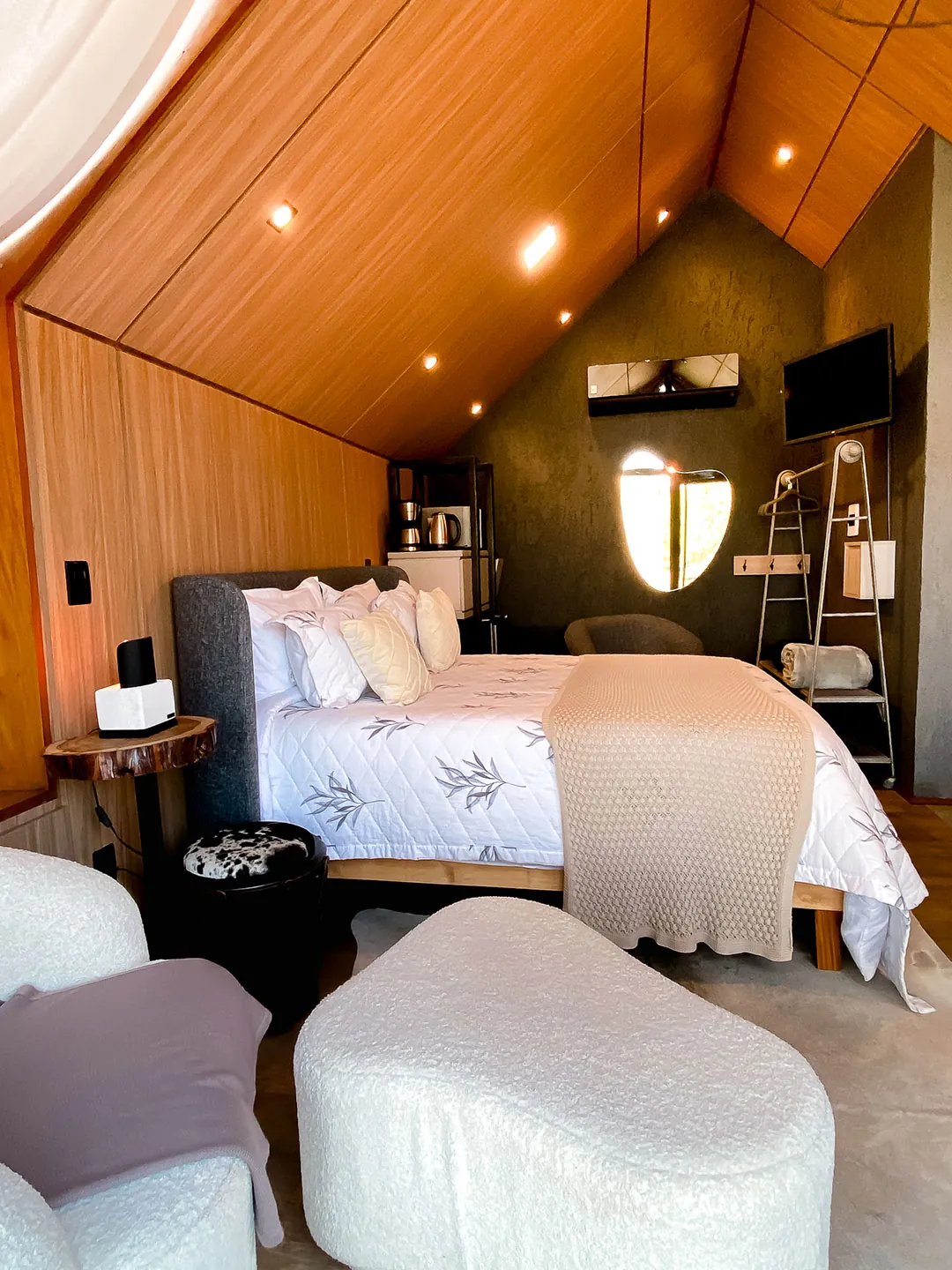Ambiente com cama de casal, pufes brancos, ar-condicionado e televisão sob o teto de madeira inclinado