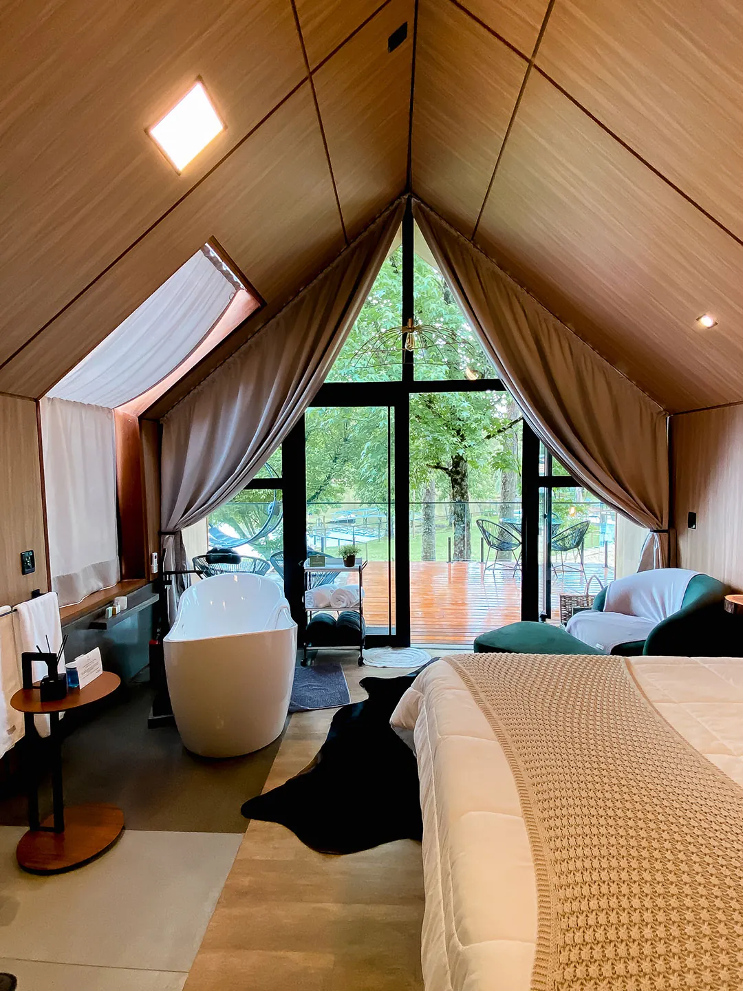 Cama de casal em primeiro plano, banheira de imersão e portas de vidro abertas para o deck molhado e árvores