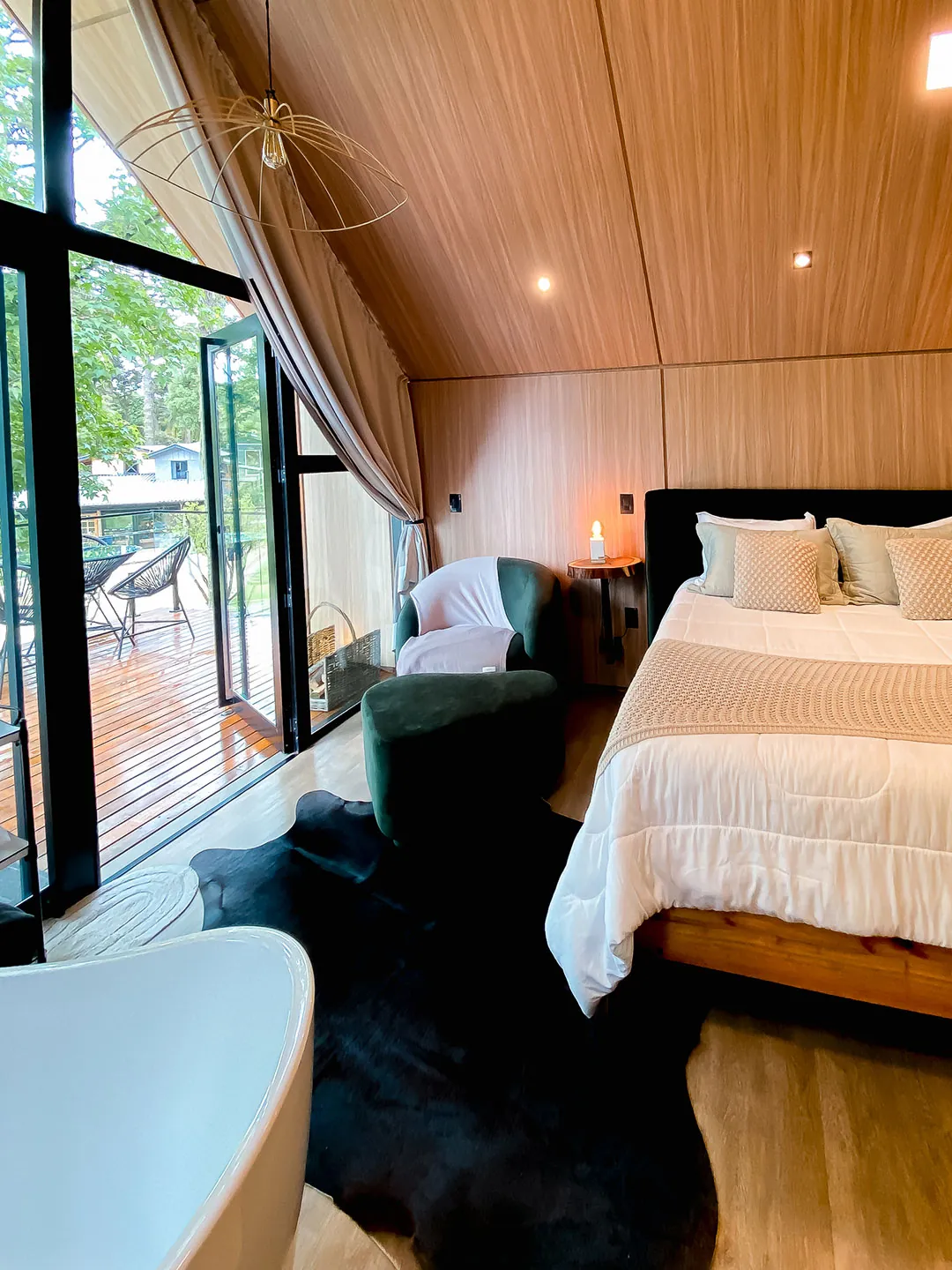 Cama de casal com cabeceira escura, poltrona verde e tapete de couro, com porta de vidro aberta para o deck