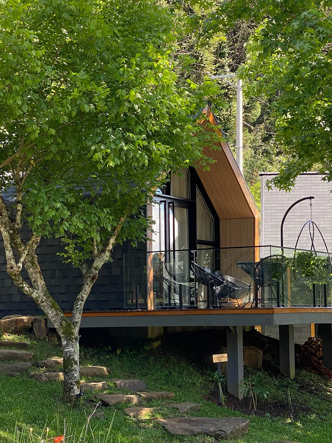Lake house sobre pilares com deck de guarda-corpo de vidro, parcialmente encoberta por uma árvore, com degraus de pedra no gramado