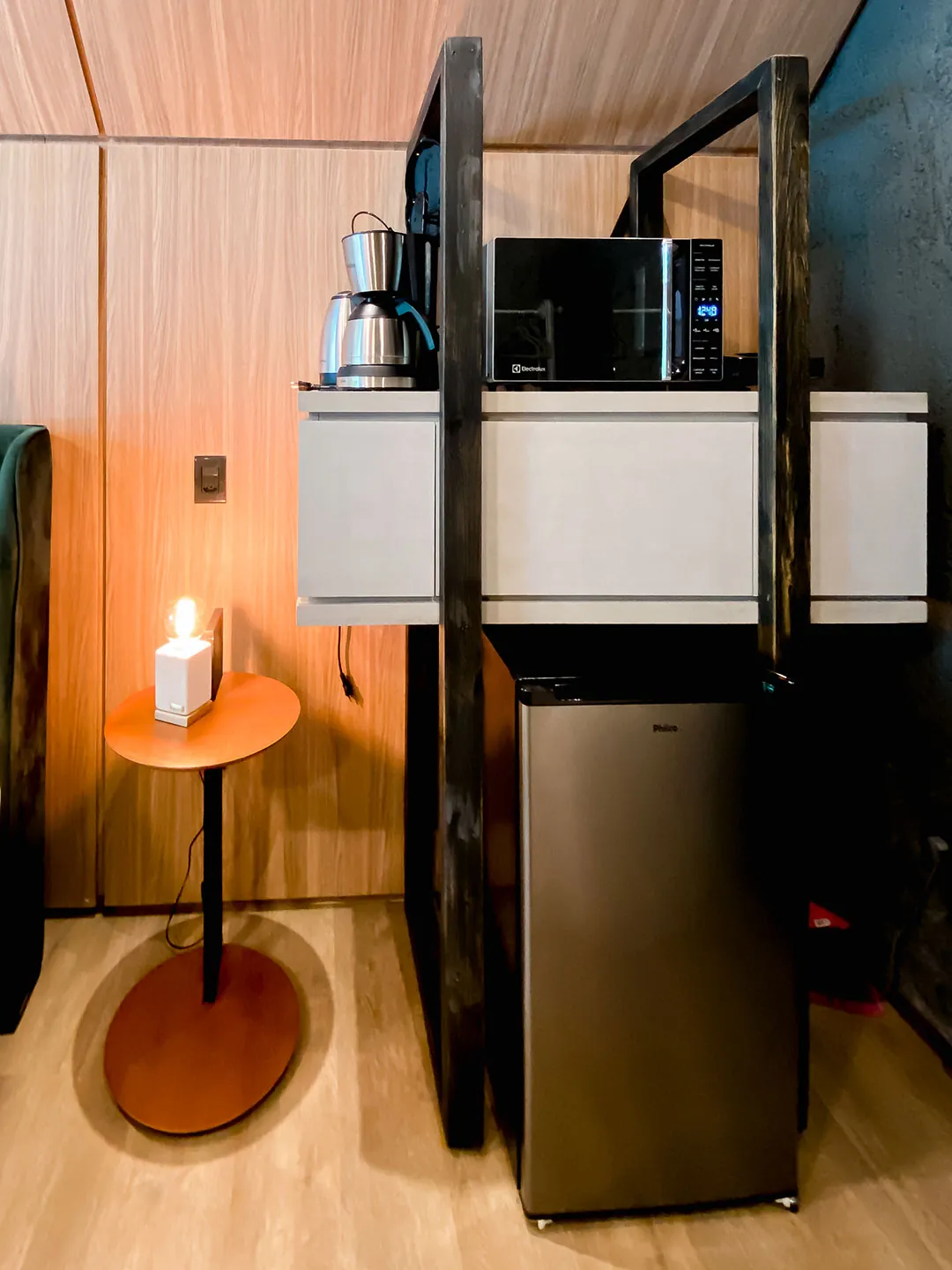 Estante metálica com cafeteira e micro-ondas sobre frigobar, ao lado de luminária de mesa