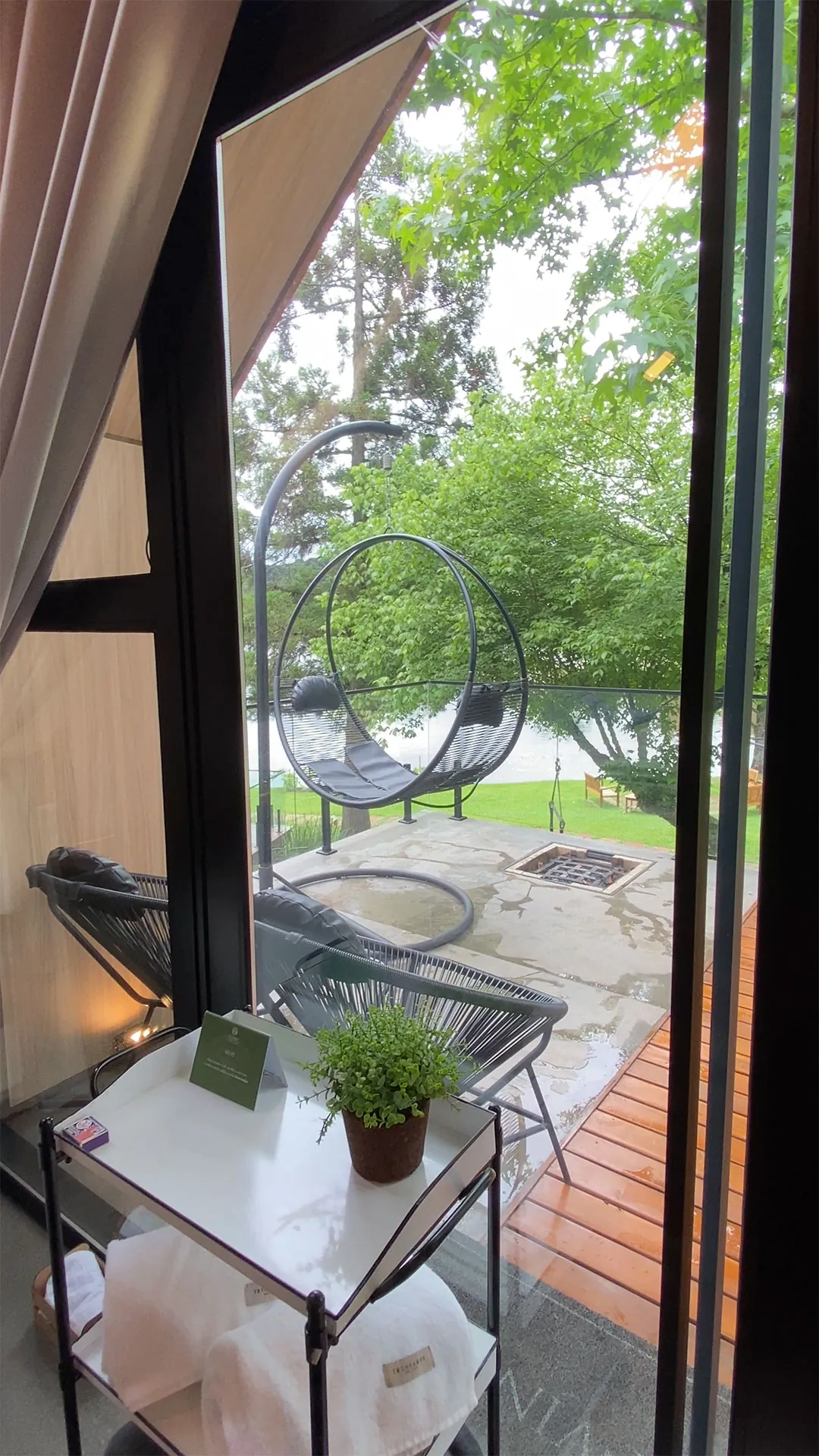 Vista pela porta de vidro do deck com poltrona suspensa e cadeira, árvores e lago ao fundo, com carrinho de toalhas em primeiro plano