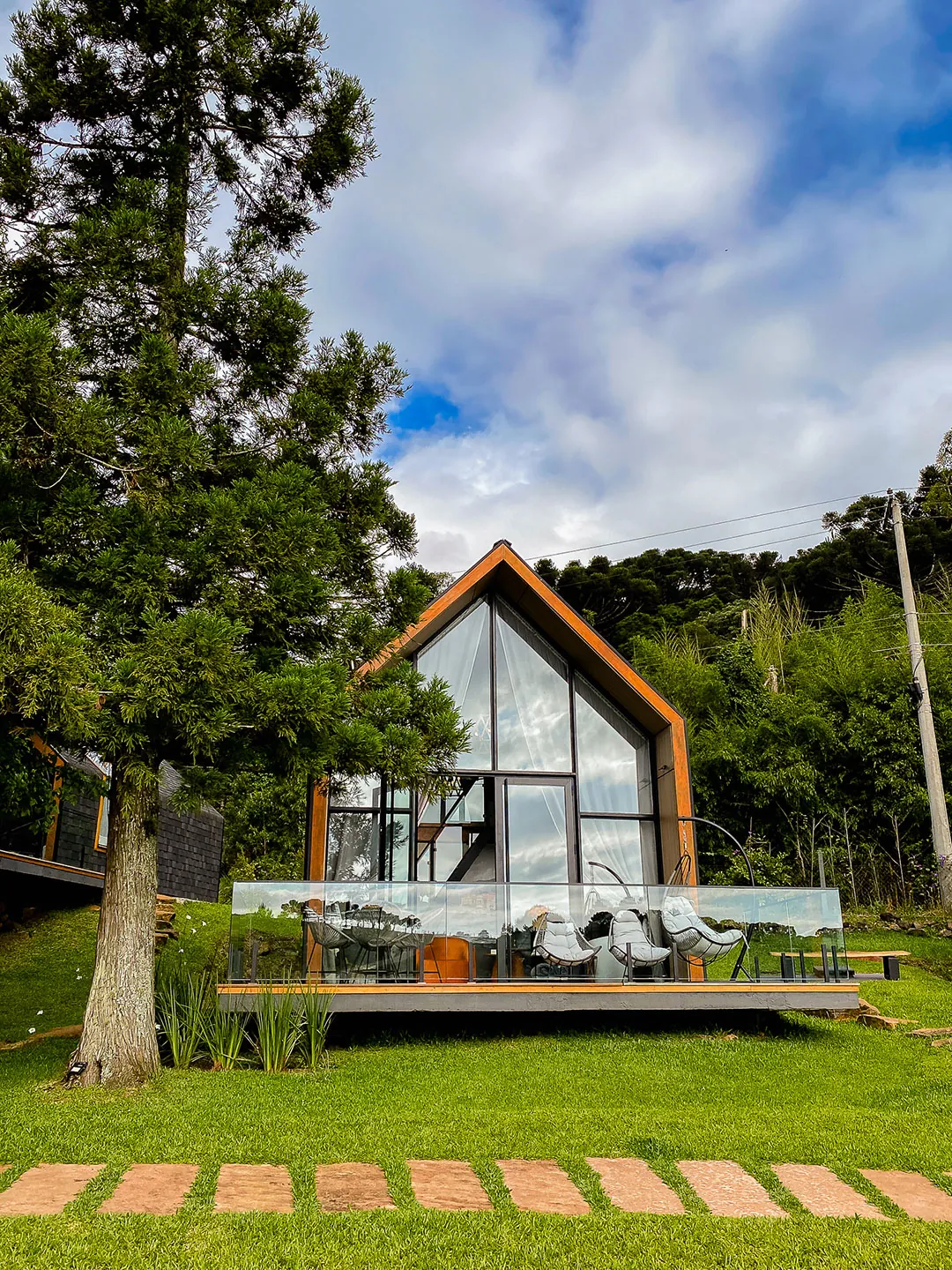 Lake house de fachada envidraçada com deck e guarda-corpo de vidro sobre gramado, com caminho de pedras e araucária à esquerda