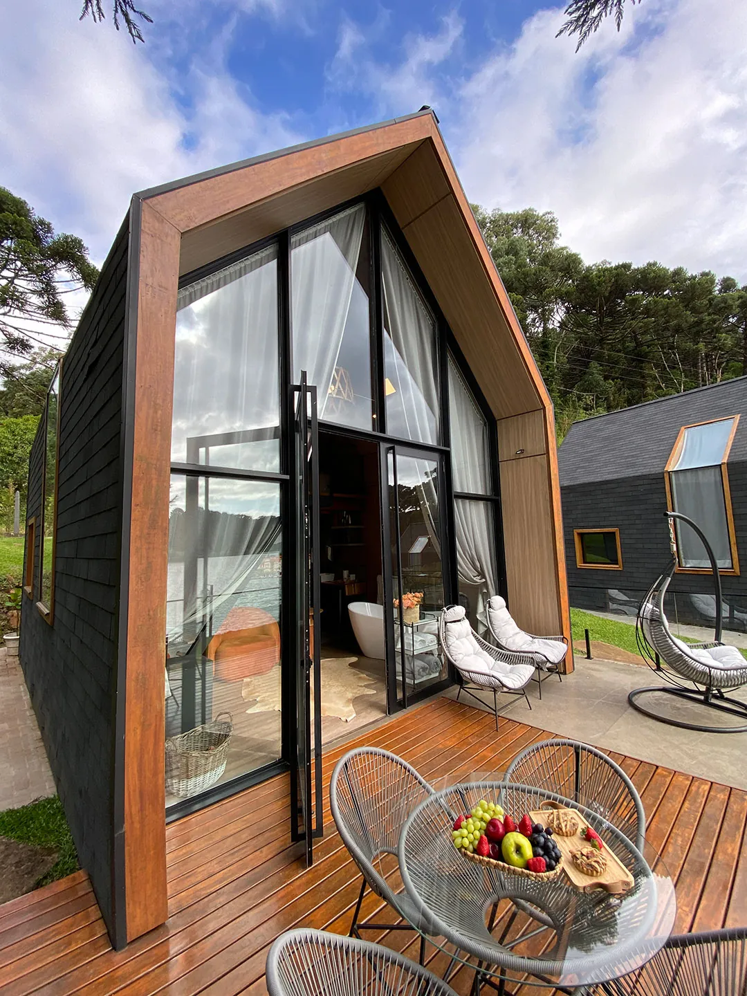 Deck de madeira com mesa de vidro, cadeiras de corda e frutas diante da porta aberta da lake house, com poltrona suspensa ao lado