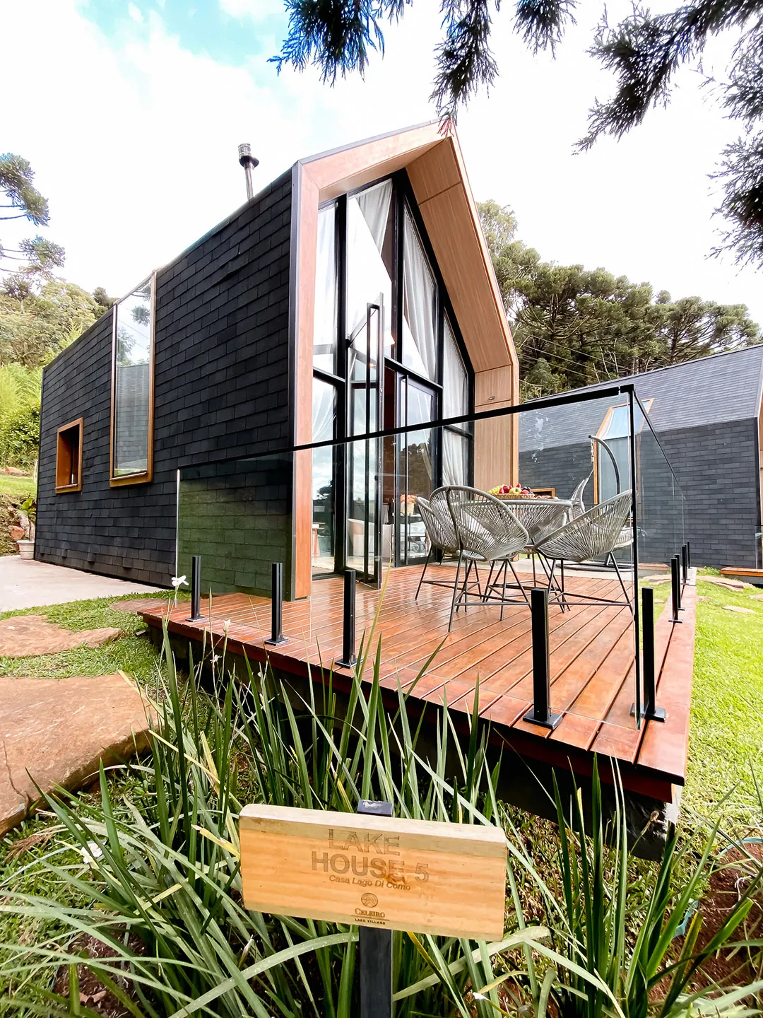 Lake house de telhas escuras com deck de madeira e guarda-corpo de vidro, com placa de identificação entre plantas em primeiro plano