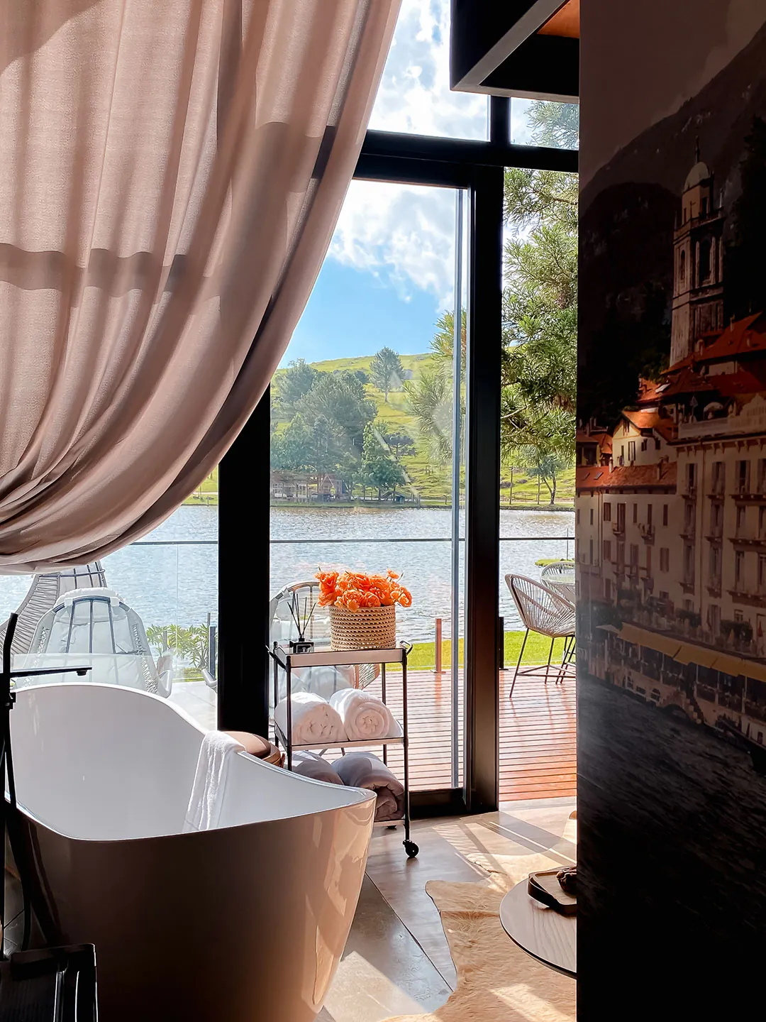 Banheira de imersão junto à porta de vidro aberta para o deck, com lago e morros ao fundo e painel fotográfico de vila na parede à direita