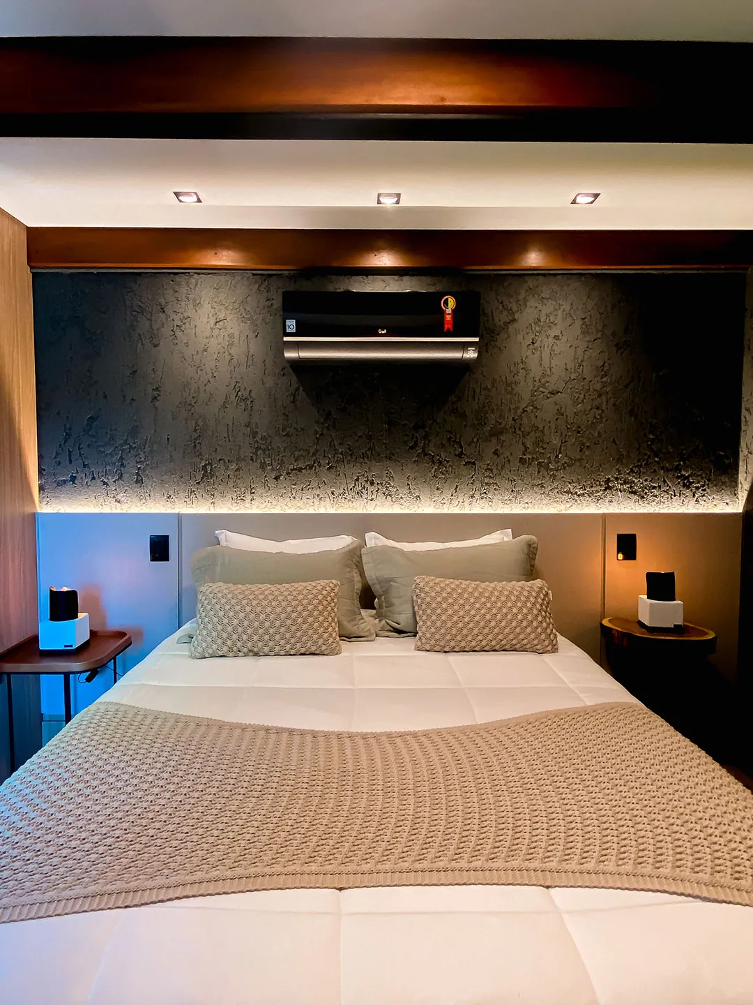 Quarto com cama de casal, almofadas verdes e tricotadas, ar-condicionado e parede texturizada escura com iluminação indireta