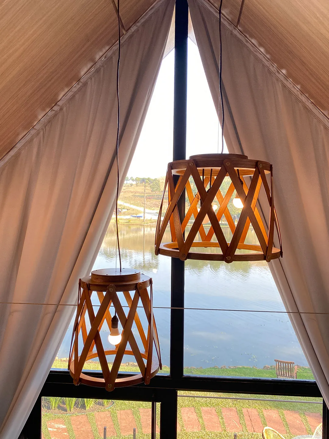Luminárias de madeira vazadas diante da janela triangular com cortinas abertas e vista para o lago