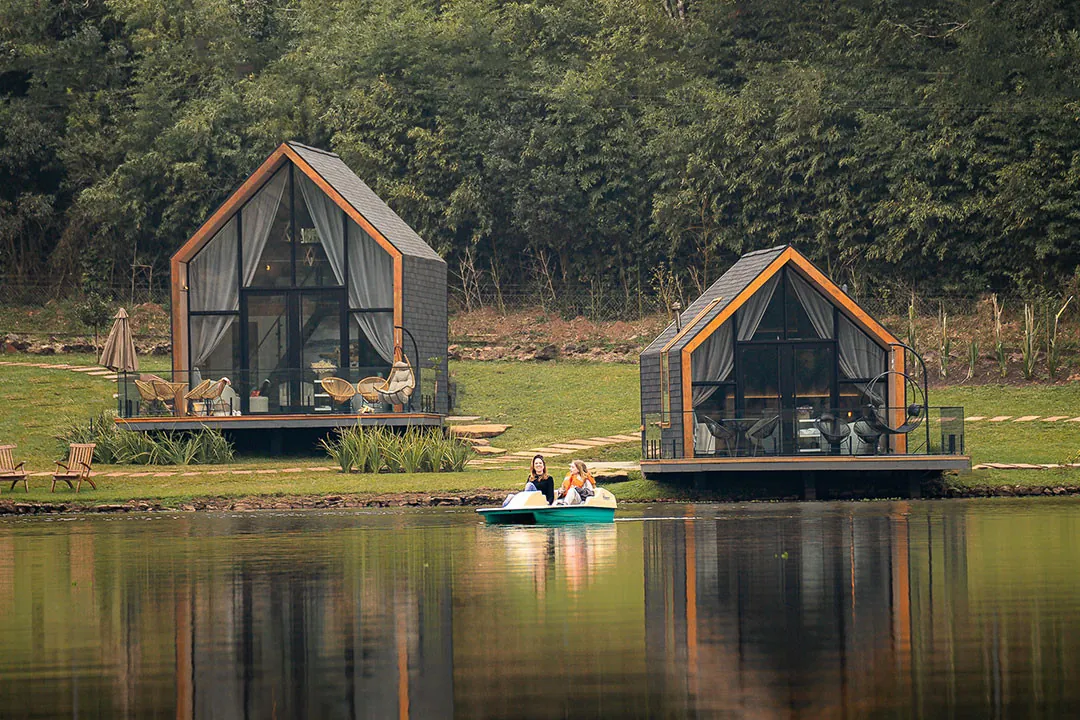 Duas lake houses com decks na margem do lago, refletidas na água, e duas pessoas em um pedalinho à frente, com mata ao fundo