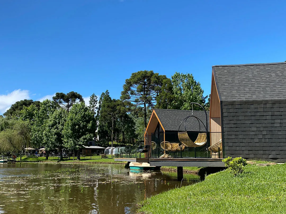Lake house com deck sobre pilares na margem do lago, poltrona suspensa e araucárias sob céu azul, ao lado de outra construção de telhas escuras