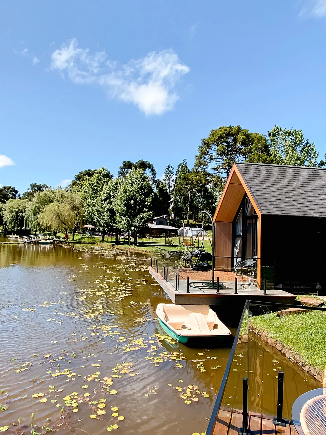 Lake house com deck sobre a água e pedalinho junto à margem, lago com plantas aquáticas e árvores ao fundo
