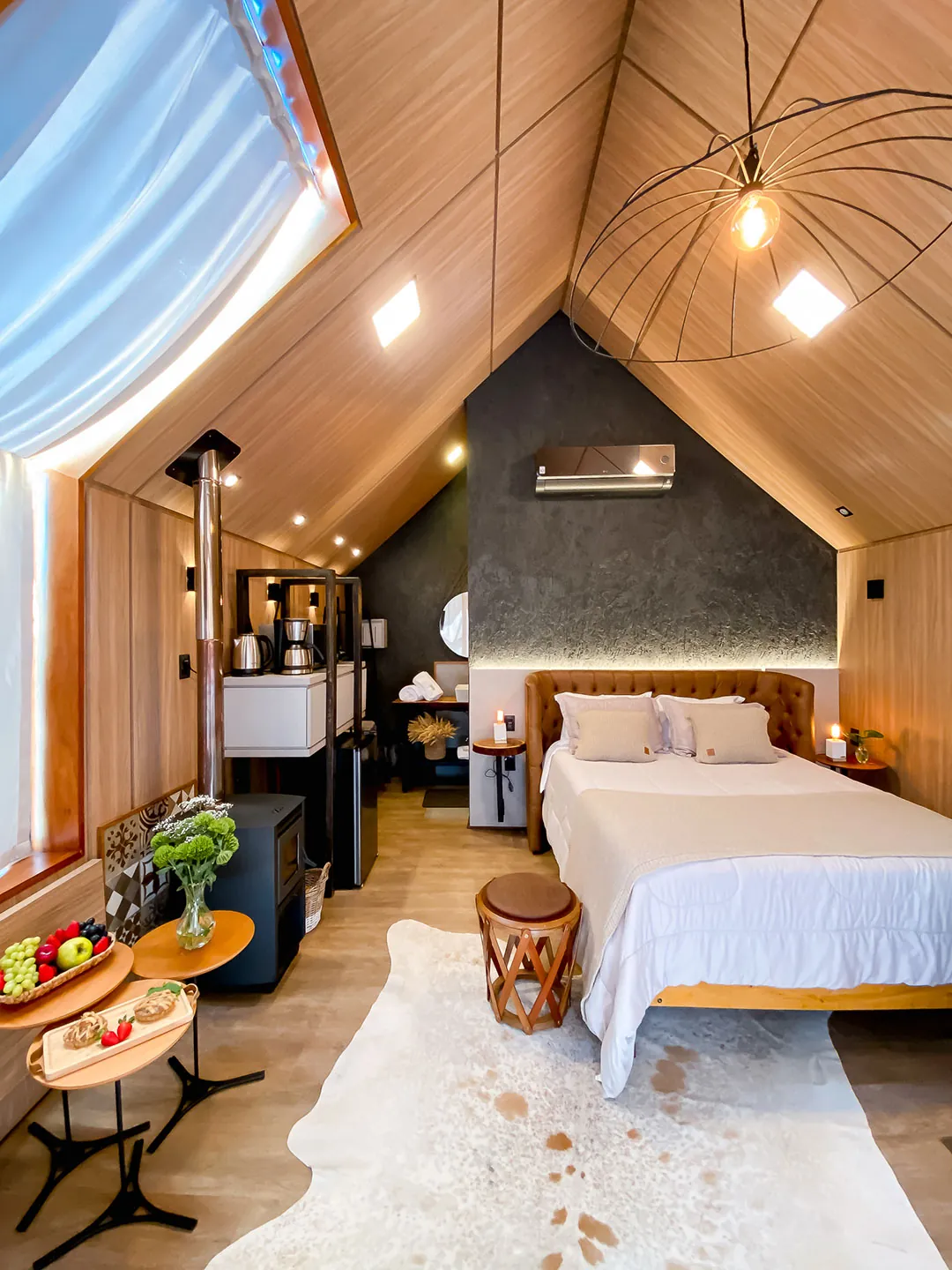 Ambiente único com cama queen de cabeceira de couro, lareira a lenha, estante com cafeteira e mesas com frutas sobre tapete de couro