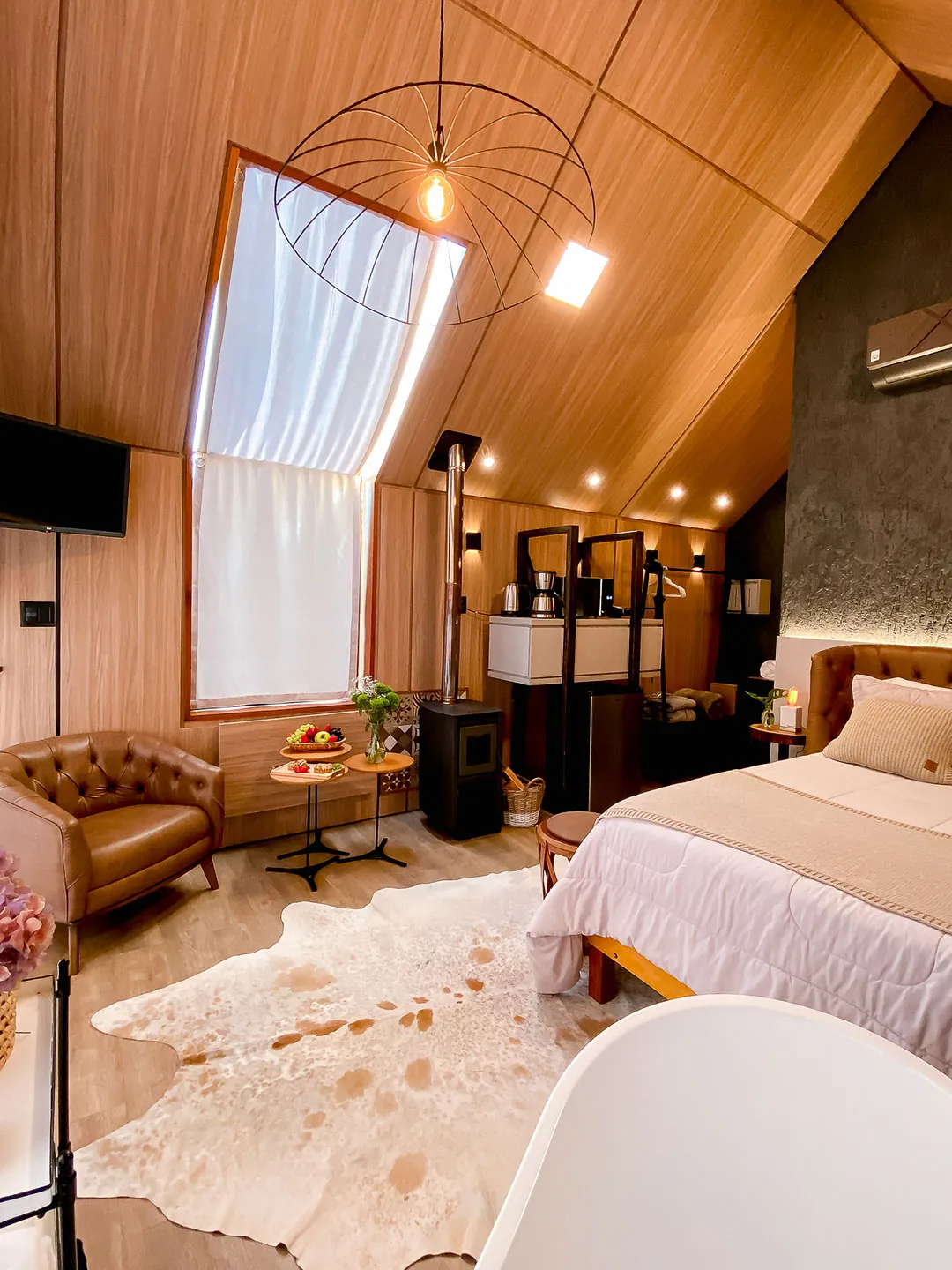 Ambiente com cama de casal, poltrona de couro, mesas com frutas, lareira a lenha e tapete de couro sob teto de madeira com lustre aramado
