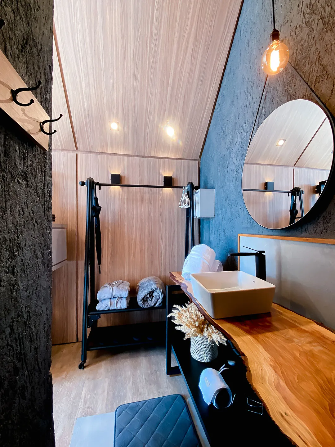 Bancada de madeira com cuba, espelho redondo, arara com roupões e luminária pendente