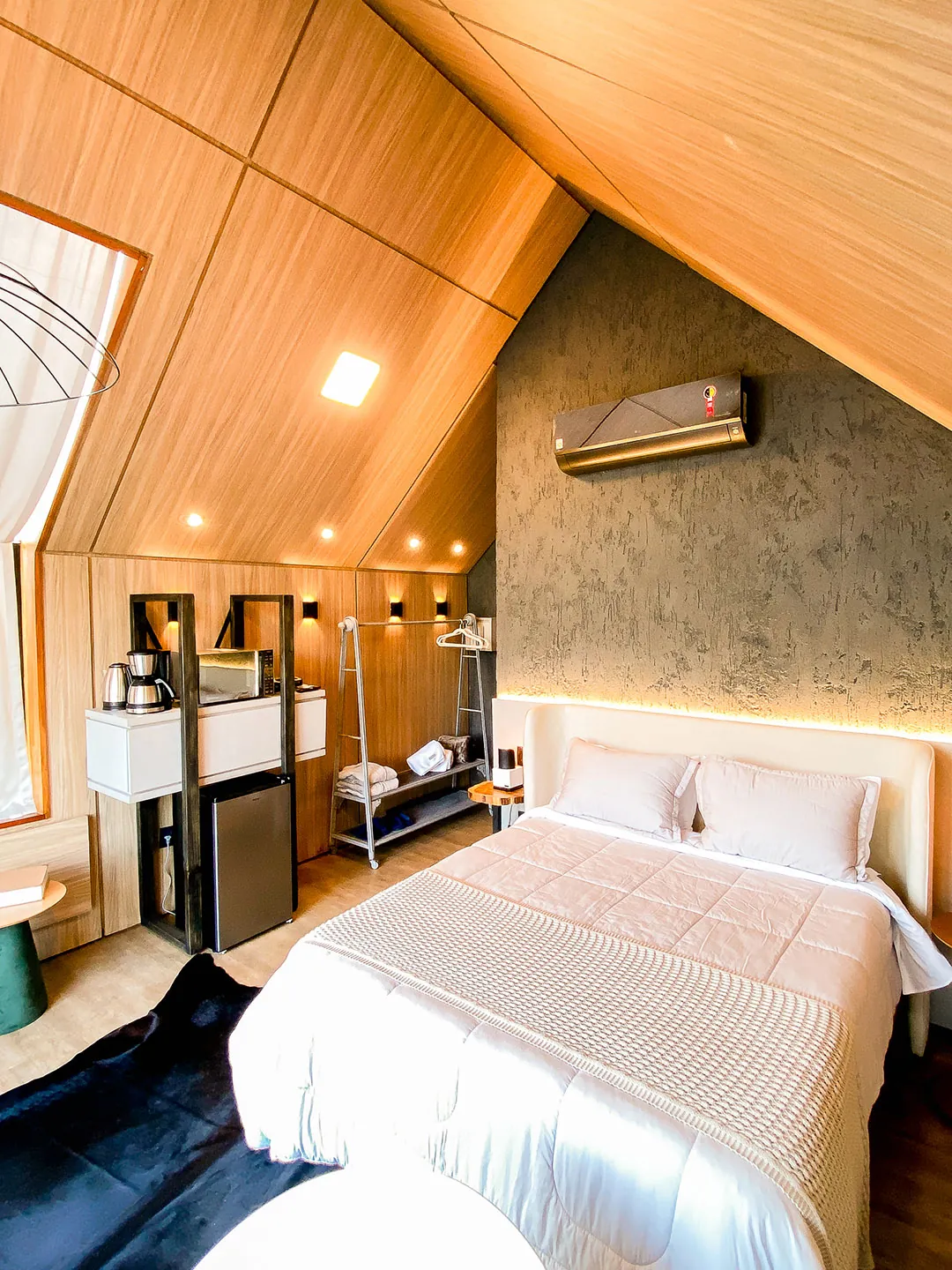 Cama queen com manta tricotada, estante com cafeteira e micro-ondas, frigobar e arara sob o teto de madeira inclinado