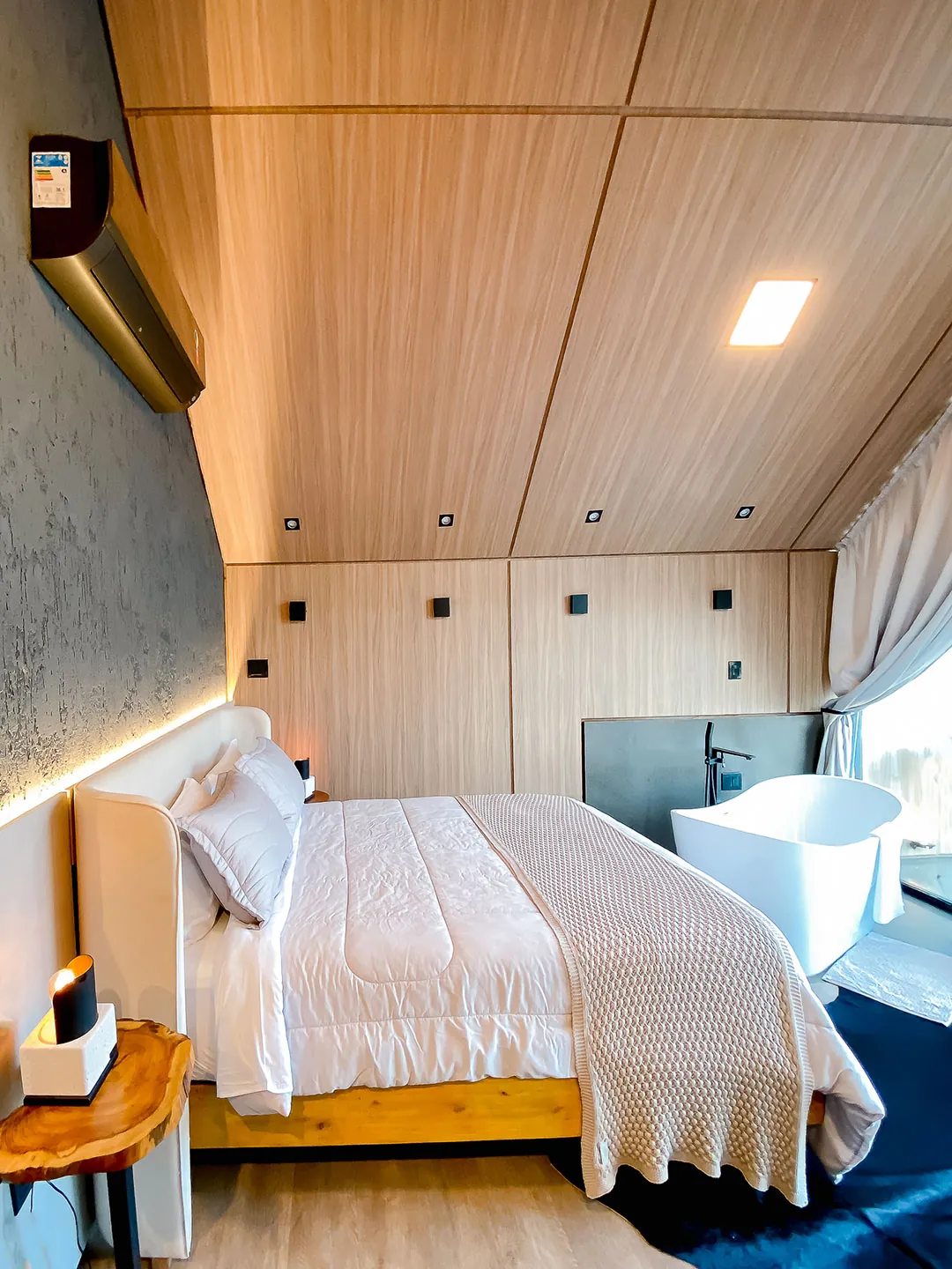 Cama de casal ao lado de banheira de imersão junto à janela, com teto de madeira e ar-condicionado