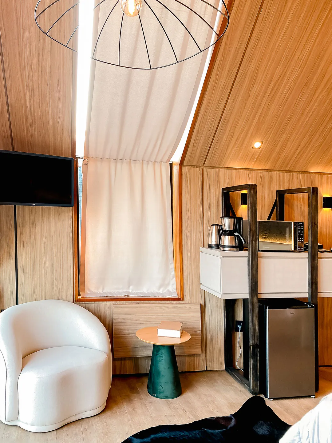 Poltrona branca, mesa lateral verde e estante com cafeteira, micro-ondas e frigobar ao lado da janela com cortina