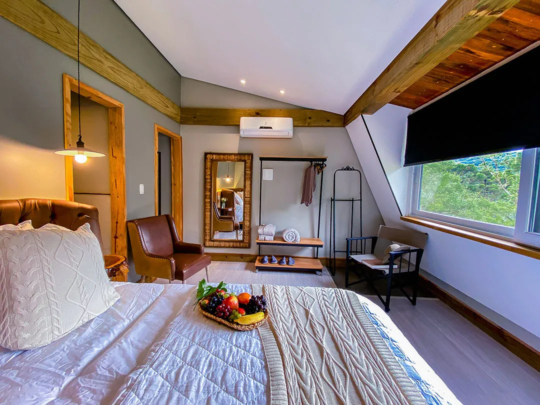 Quarto de teto inclinado com cama de casal, almofada de tricô e cesta de frutas; poltrona de couro, espelho com moldura de madeira, arara e janela ampla com persiana escura.
