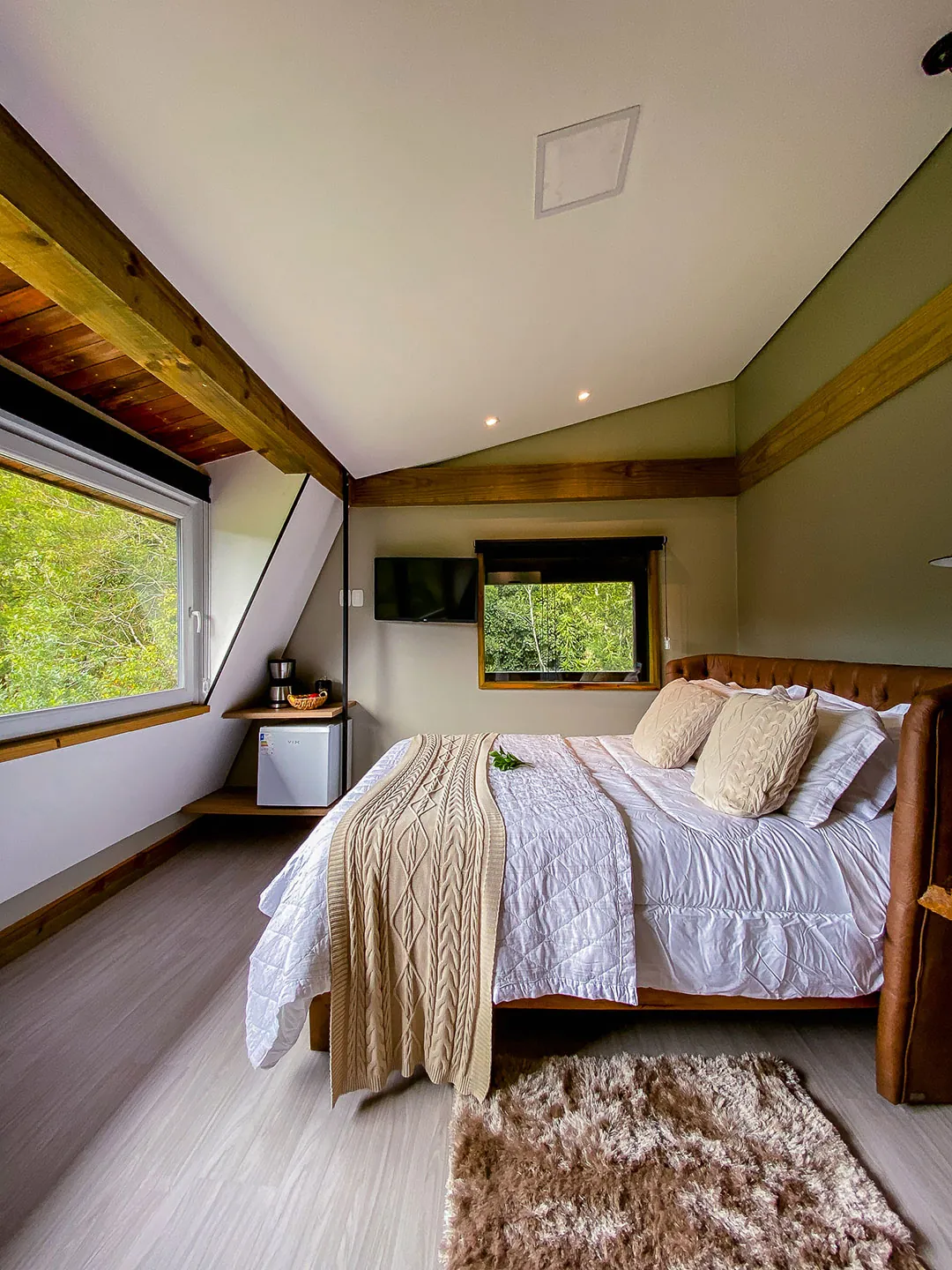 Cama de casal com cabeceira de couro, manta de tricô bege e tapete felpudo; ao lado, cafeteira e frigobar; janelas com vista para árvores.