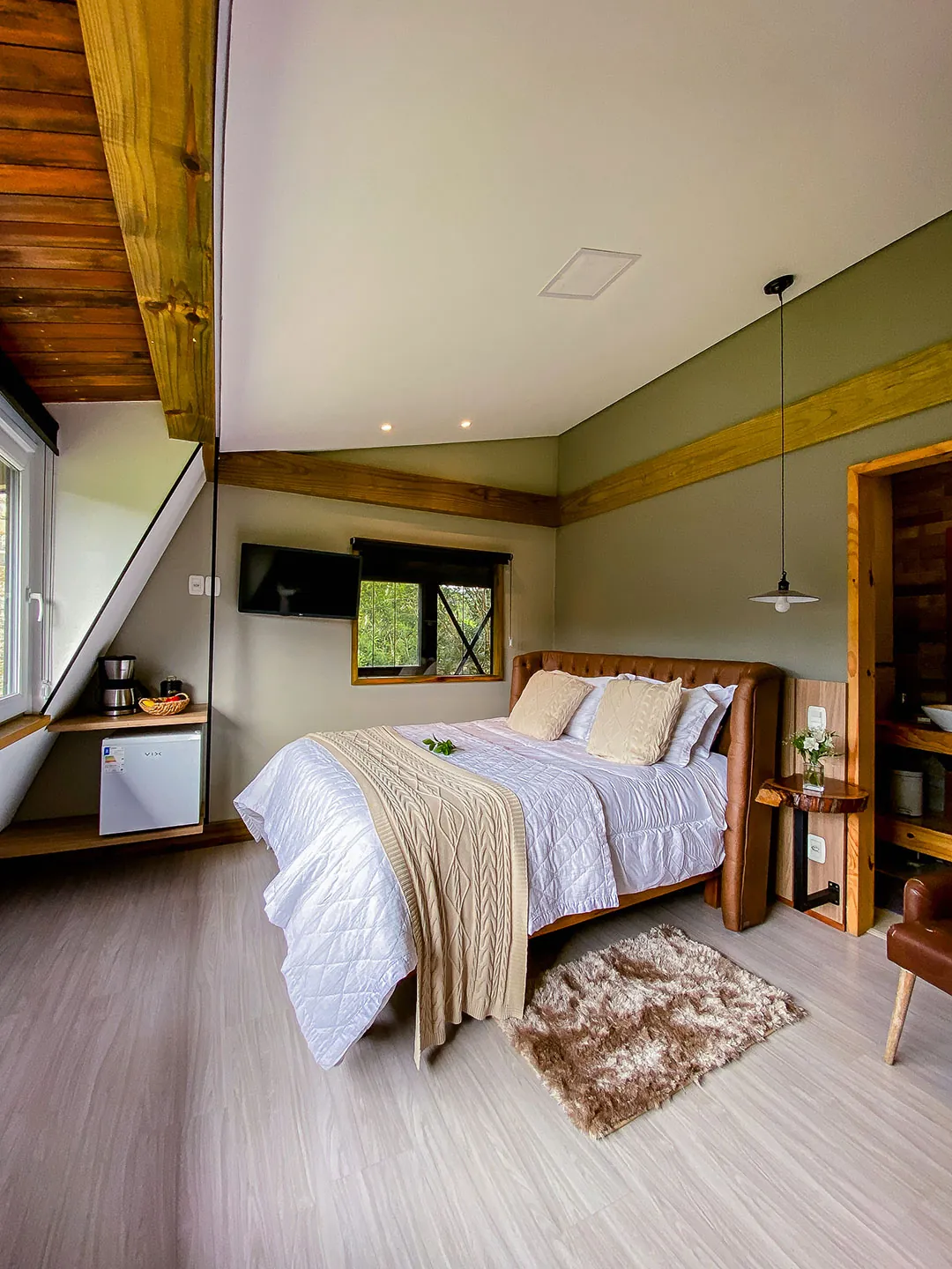 Cama de casal com manta de tricô bege, tapete felpudo, mesa lateral com flores e porta de madeira à direita; teto inclinado com viga de madeira.