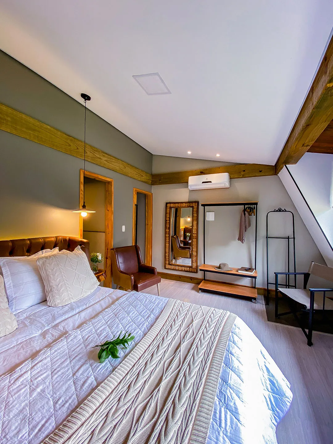 Quarto visto da cama: poltrona de couro, espelho com moldura de madeira, arara, ar-condicionado e janela inclinada; ramo verde sobre a manta.