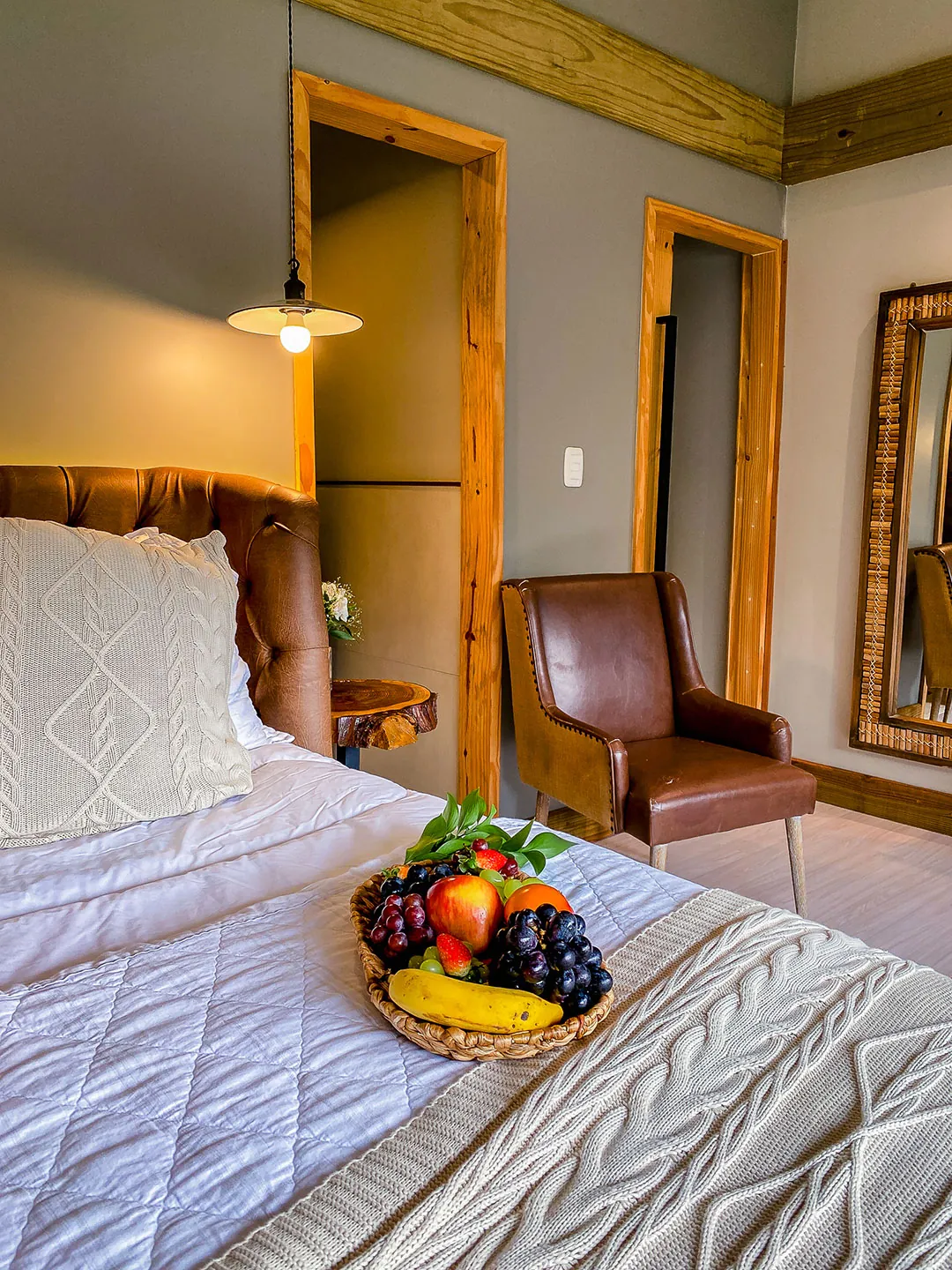 Cesta com frutas sobre a cama com manta de tricô; ao fundo, poltrona de couro, luminária pendente, portas de madeira e espelho.