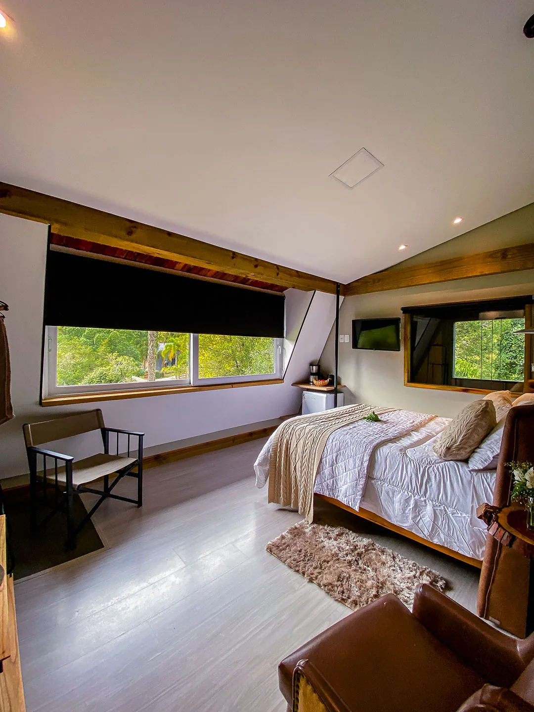 Quarto com banco de madeira e lona, cama de casal com manta, tapete felpudo e janela ampla com persiana escura e vista para árvores; poltrona em primeiro plano.