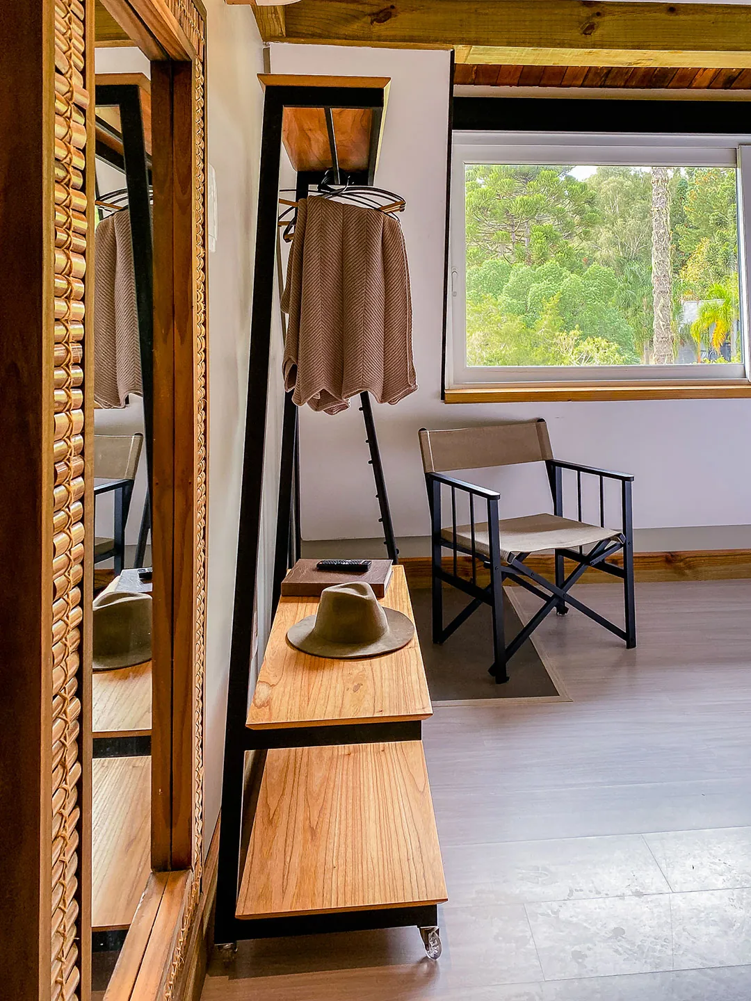 Arara de metal com chapéu e peça de roupa, cadeira dobrável e janela com vista para araucárias; espelho de moldura de madeira em primeiro plano.