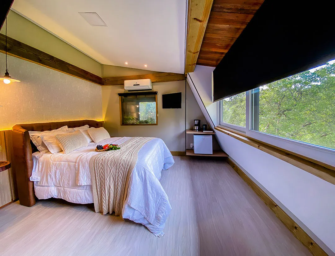 Quarto de teto inclinado com cama de casal, cabeceira de couro, manta de tricô e cesta de frutas; janela ampla com persiana escura e vista para árvores; frigobar e cafeteira ao fundo.