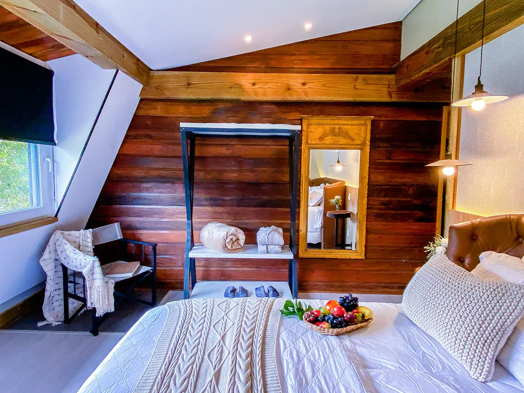 Cama com manta de tricô e cesta de frutas em frente a parede de tábuas de madeira escura com estante de metal, toalhas enroladas, pantufas, espelho de moldura dourada e cadeira com manta.
