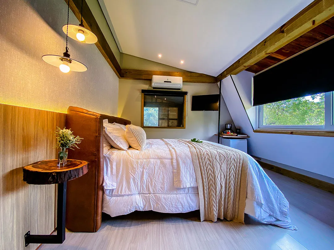 Cama de casal vista de lado com cabeceira de couro, mesa lateral de tronco com flores, luminárias pendentes acesas e janela ampla com vista para árvores.