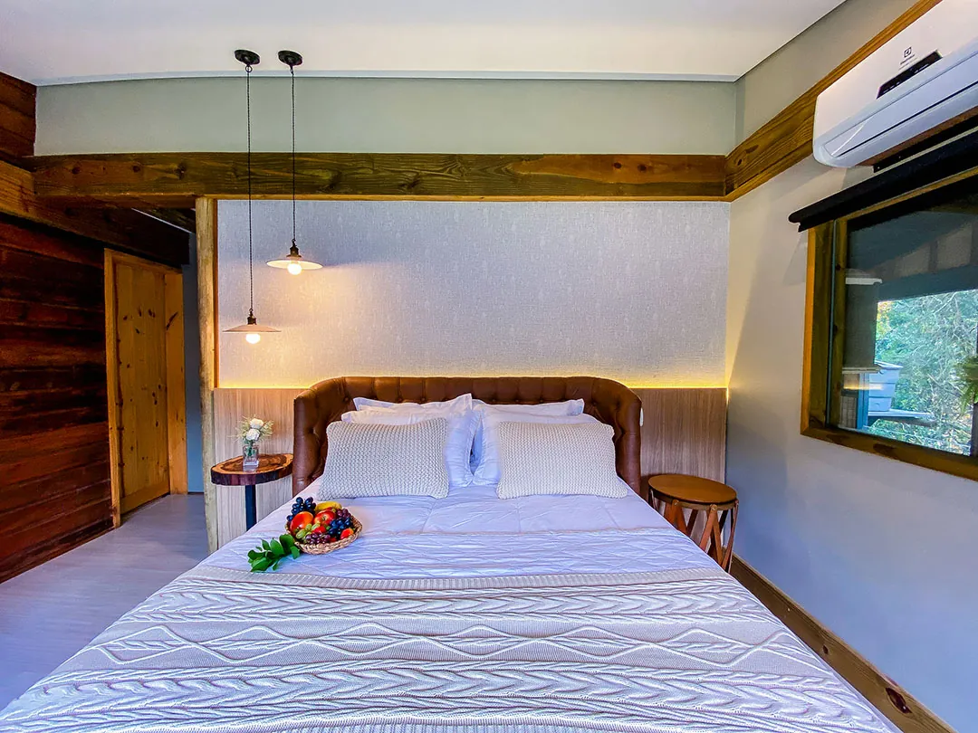 Vista frontal da cama de casal com cesta de frutas, luminárias pendentes, parede de tábuas de madeira à esquerda e janela à direita.