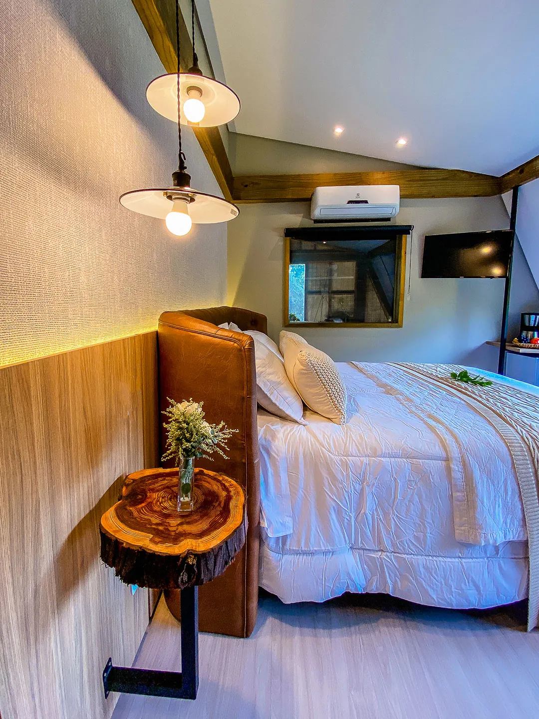 Cama com cabeceira de couro vista de lado, mesa lateral de tronco com flores e luminária pendente acesa; ao fundo, ar-condicionado, TV e janela pequena.
