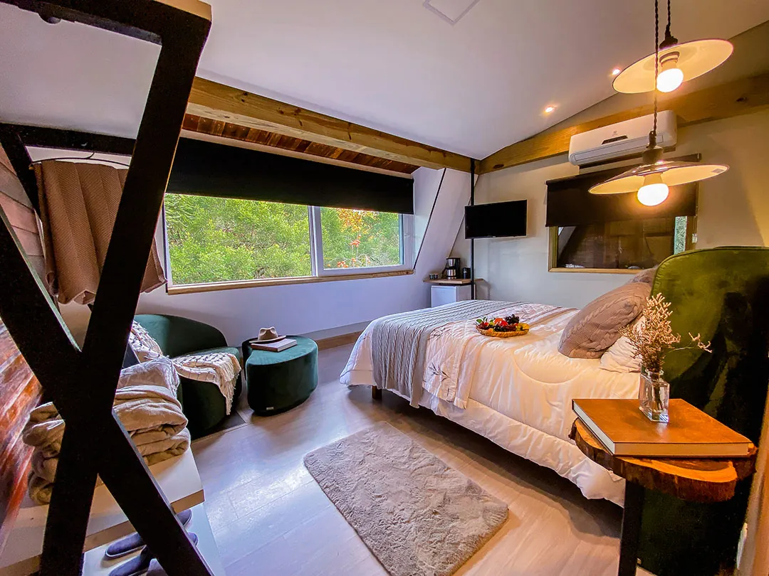 Vista ampla do quarto com cama de casal, bandeja sobre a manta, tapete, poltronas e puff verdes, mesa lateral com flores, luminárias acesas e janela com vista para árvores; estrutura de metal em X em primeiro plano.