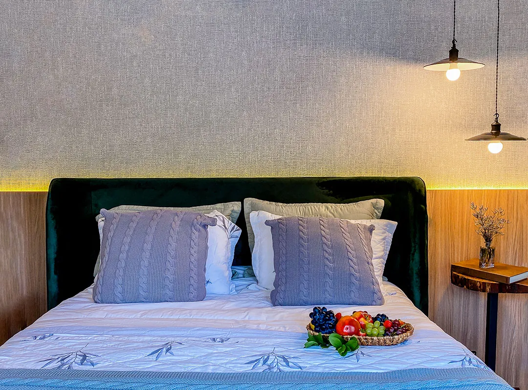 Cama com cabeceira de veludo verde, almofadas de tricô azul-acinzentadas, cesta de frutas, mesa lateral de madeira e luminárias pendentes.