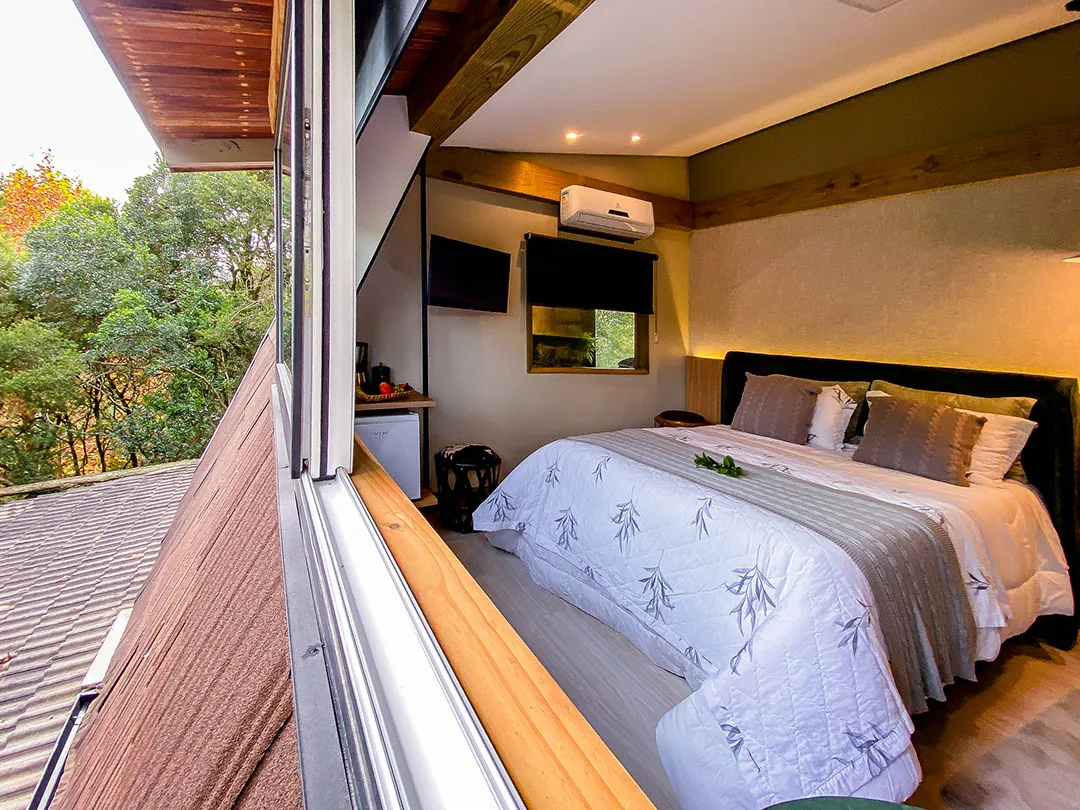 Quarto visto de fora pela janela aberta: cama de casal com roupa de cama estampada, TV, ar-condicionado e cafeteira; à esquerda, telhado e copas de árvores.