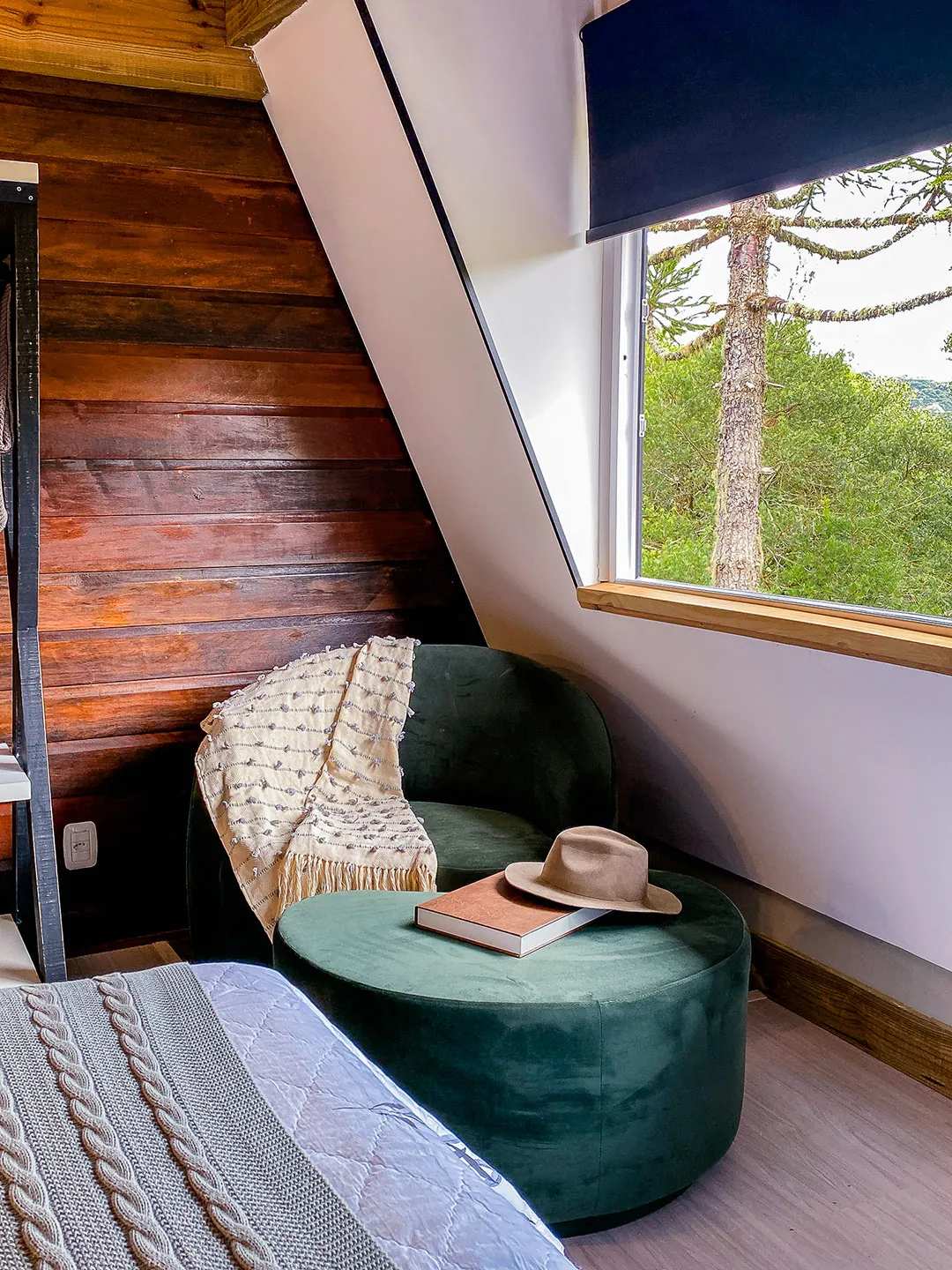 Poltrona e puff de veludo verde com chapéu e livro, junto a parede de tábuas de madeira e janela inclinada com vista para araucária.