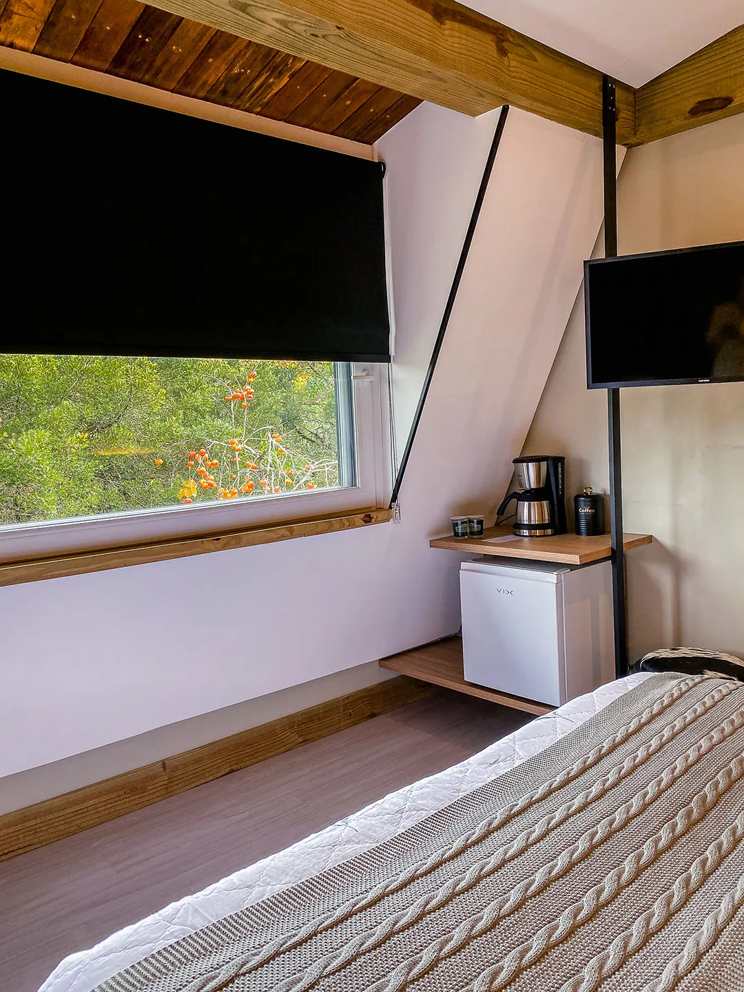 Janela ampla com persiana escura e vista para árvores, cafeteira e frigobar em prateleira de madeira, TV na parede; pé da cama em primeiro plano.
