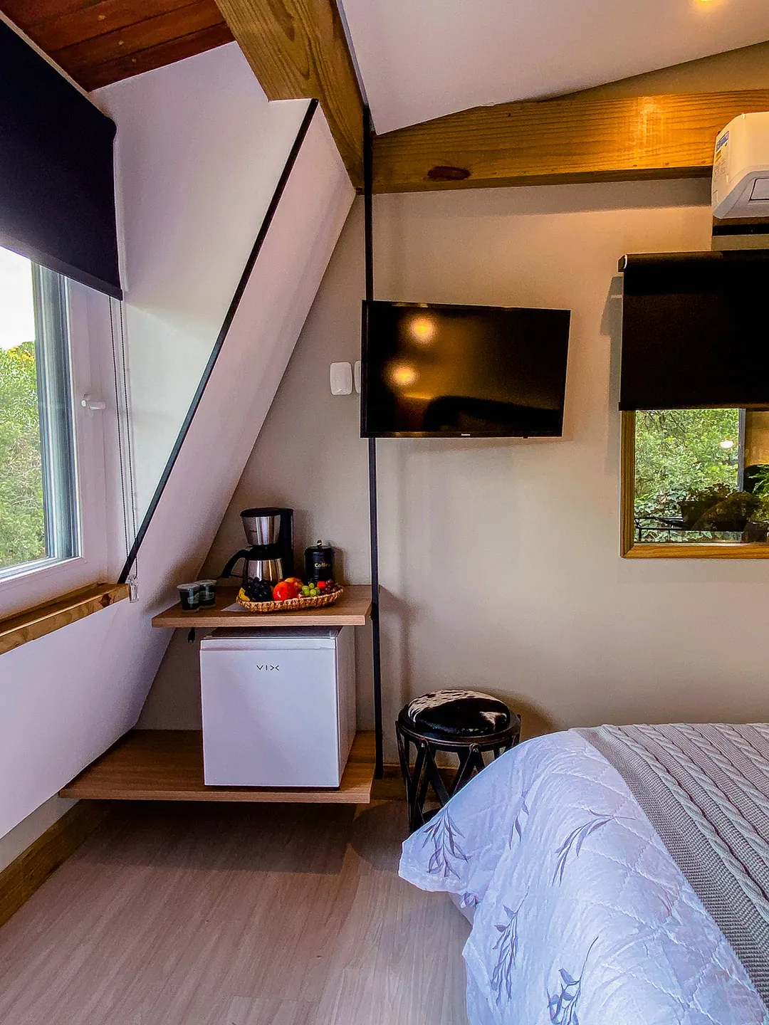 Canto com TV na parede, cafeteira, cesta e frigobar em prateleira de madeira, banqueta e janela inclinada; cama em primeiro plano.