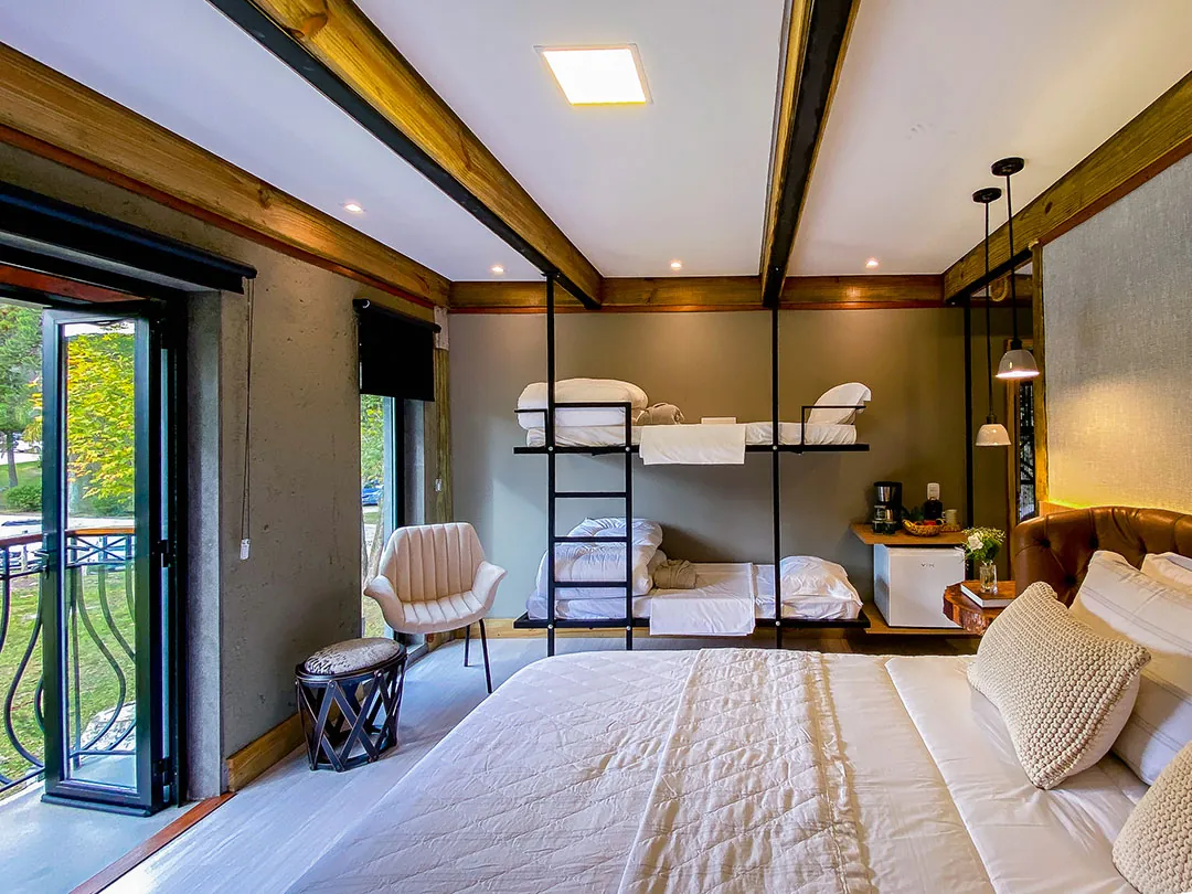 Quarto com cama de casal em primeiro plano, beliche de metal com roupa de cama branca, poltrona, banqueta e porta de vidro aberta para sacada com gradil; teto branco com vigas de madeira.