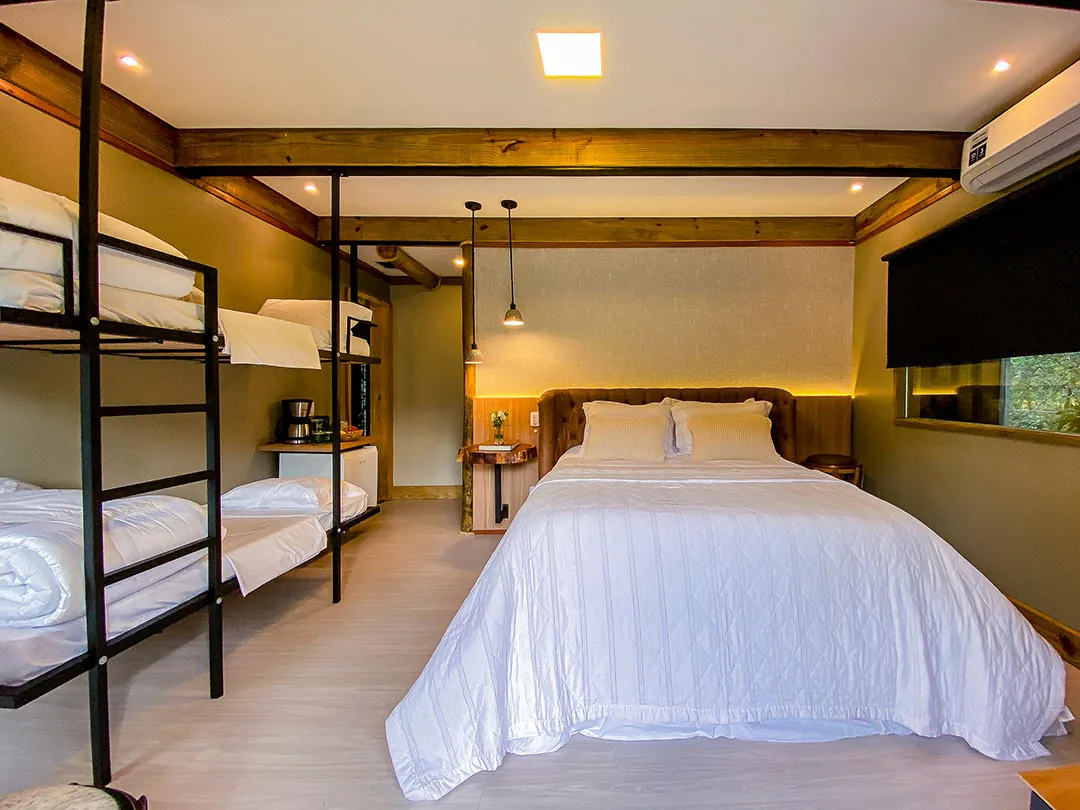 Vista frontal do quarto: cama de casal com roupa de cama branca, beliches de metal à esquerda, cafeteira e frigobar ao fundo e janela com persiana escura à direita.