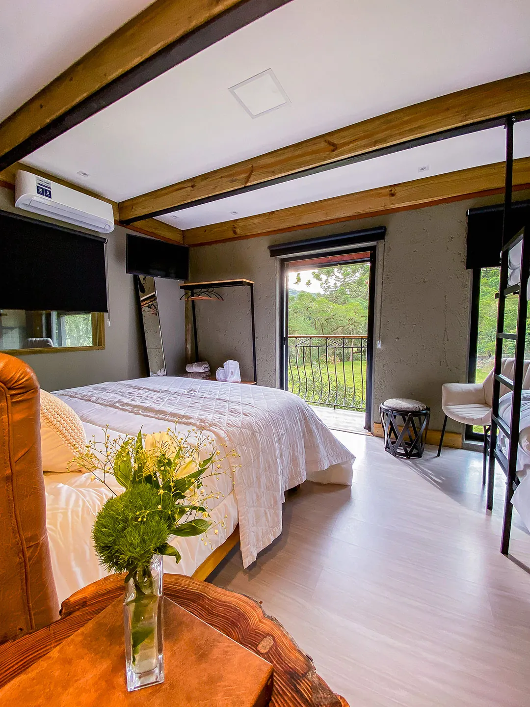 Cama de casal vista da cabeceira, com mesa lateral de tronco e vaso de flores em primeiro plano; ao fundo, porta aberta para a sacada, arara e poltrona.
